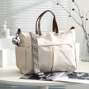 Puede incluir: Bolso tote de color crema con asas de cuero marrón y una correa para el hombro estampada. El bolso tiene dos bolsillos exteriores y un compartimento principal espacioso. Una revista y un reloj están sobre la mesa.