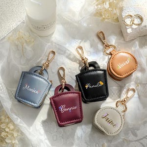 Peut inclure: Plusieurs porte-clés personnalisés de couleurs variées, dont bleu, bordeaux, noir, beige et blanc. Chaque porte-clés est doté d'un fermoir doré et d'un nom inscrit en écriture délicate. Certains porte-clés ont des motifs floraux.