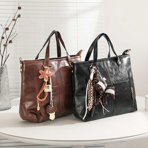 Può includere: Due borse tote in pelle, una marrone e una nera, sono affiancate. La borsa marrone ha un ciondolo a forma di stella e un ciondolo a forma di fungo. La borsa nera ha una tasca e nappe decorative. Entrambe hanno manici superiori.