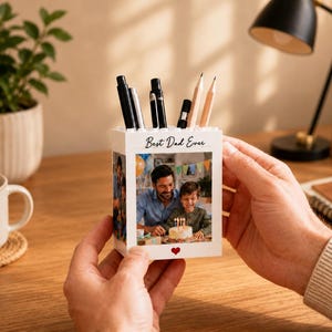 May include: A white pen holder with a photo of a father and child, and the text "Best Dad Ever." The holder contains pens and pencils. A small red heart is visible below the photo. The pen holder is held in a person's hands.