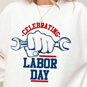 Könnte beinhalten: Weißes Sweatshirt mit einer Grafik zum Tag der Arbeit. Das Design zeigt eine Hand, die einen Schraubenschlüssel hält, mit den Worten "CELEBRATING LABOR DAY" in Rot und Blau. Ein lässiges, bequemes Oberteil.