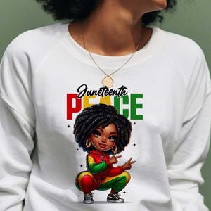 Puede incluir: Sudadera blanca con una colorida ilustración de una niña negra con un atuendo temático del Juneteenth. Las palabras "Juneteenth Peace" están impresas encima de la ilustración en rojo, verde y negro.