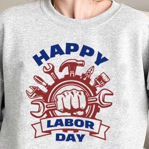 Puede incluir: Una sudadera gris claro con un diseño gráfico para el Día del Trabajo. El diseño presenta un engranaje rojo con herramientas, un puño que sostiene una llave inglesa y las palabras "HAPPY LABOR DAY" en azul y rojo.