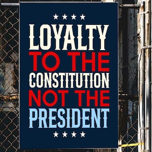 May include: A rectangular navy blue banner with white and red text that reads "LOYALTY TO THE CONSTITUTION NOT THE PRESIDENT." The banner is decorated with white stars and hangs on a chain-link fence.
