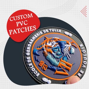 Może przedstawiać: Niestandardowa naszywka PVC ze szczegółowym wzorem przedstawiającym stworzenie podobne do tygrysa w odcieniach niebieskiego, pomarańczowego i białego. Naszywka ma napis "ECOLE DE GENDARMERIE DE TULLE" i "RE VOULOIR" na obrzeżu. Czerwone koło z napisem "CUSTOM PVC PATCHES" znajduje się w lewym górnym rogu.