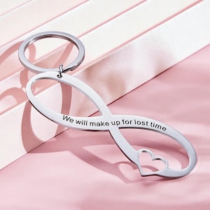 May include: A silver-colored infinity symbol keychain with a heart cutout and a ring. The keychain has the engraved text: "We will make up for lost time". The keychain is on a pink surface.