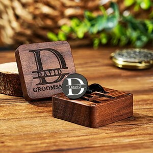 May include: A wooden box and a cufflink with the name "David" engraved. The box is square and has the word "Groomsmen" engraved. The cufflink is black and silver. The background is a wooden surface with greenery.
