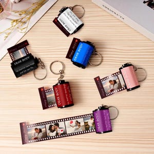 May include: A selection of film roll keyrings in black, blue, red, pink, and purple. Each keyring displays a photo strip with images and the text "DEAR BODY" and "Happy 2023". The keyrings are arranged on a light wooden surface.