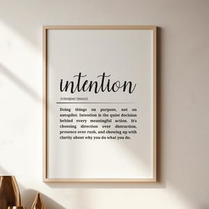 Könnte beinhalten: Ein gerahmter Druck mit dem Wort „Intention“ und seiner Definition. Drei goldene Vasen stehen auf einem Holzregal. Rechts vom Druck befindet sich eine Topfpflanze. Der Raum hat einen Holzboden und ein Fenster.