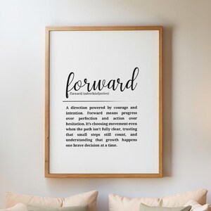 Puede incluir: Un cuadro enmarcado con la palabra "forward" en escritura negra y una definición. El cuadro está colgado sobre un sofá de mimbre con cojines beige y verdes. Una manta y una alfombra de color crema completan la escena de un salón acogedor.