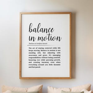 May include: A living room scene featuring a wooden framed print with the words "balance in motion". A woven sofa with beige cushions and a cream throw is in front of the print. Decorative plants and a rug complete the cozy interior.