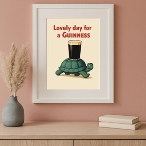 Puede incluir: Impresi&oacute;n enmarcada con una tortuga de dibujos animados que lleva una pinta de Guinness. El texto rojo dice "Lovely day for a Guinness". La imagen tiene un fondo crema y est&aacute; en un marco blanco.