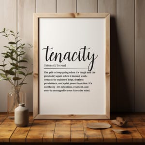 May include: A framed print featuring the word "tenacity" and its definition. The print is in a light brown wooden frame. The definition describes grit, persistence, and resilience. The print is on a wooden surface.