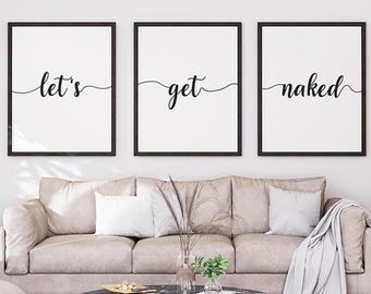 May include: Three framed wall art prints with black frames. Each print has a white background with black cursive text: "let's", "get", and "naked". The prints are displayed above a beige sofa with decorative pillows and a round coffee table.