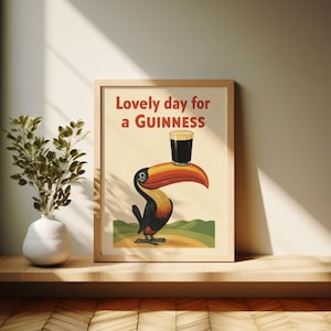 May include: Framed print featuring a cartoon toucan with a large orange beak, holding a pint of Guinness. The text "Lovely day for a Guinness" is displayed in red. The artwork is set against a beige background.