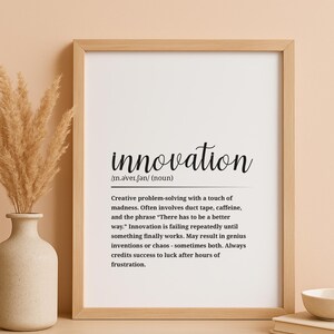 May include: A framed print with the definition of "innovation" in black text. The definition includes phrases like "duct tape, caffeine" and "genius inventions or chaos." The frame is light brown, and the background is a neutral beige.