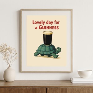 Puede incluir: Impresi&oacute;n enmarcada con una tortuga de dibujos animados que lleva una pinta de Guinness. El texto "Lovely day for a Guinness" se muestra en rojo y marr&oacute;n. El fondo es de color beige claro. Ilustraci&oacute;n humor&iacute;stica.