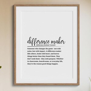 May include: A framed print with the words "difference maker" in a stylish font. The definition of "difference maker" is written below the title, describing someone who makes a positive impact. The frame is a light brown color.