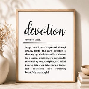 May include: A framed wall art print featuring the word devotion in a large black script font, followed by its phonetic pronunciation and a dictionary-style definition describing deep commitment, loyalty, focus, and care in black text.