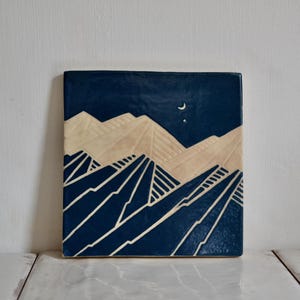 May include: A square ceramic tile featuring a deep blue background with a carved mountain landscape in cream. The design includes layered mountain peaks, a small crescent moon, and a single star in the upper section, rendered in a flat, graphic style.
