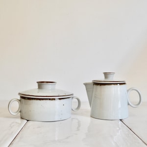 May include: Two-piece ceramic serving set. The set includes a lidded casserole dish with two handles and a teapot with a spout and handle. Both pieces are off-white with brown trim.