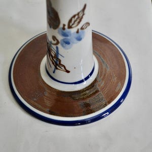 Puede incluir: Un portavelas de cer&aacute;mica blanca con un dise&ntilde;o floral marr&oacute;n y azul. La base del portavelas es un c&iacute;rculo marr&oacute;n con un borde azul.