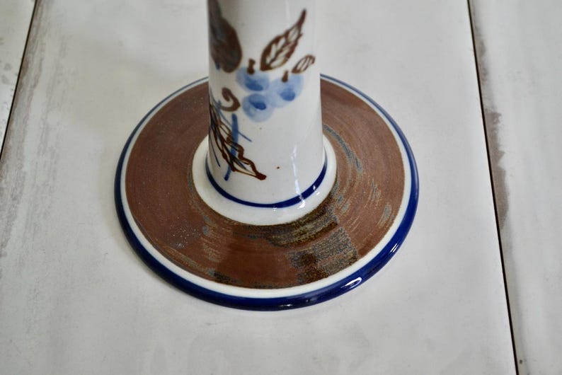 Puede incluir: Un portavelas de cer&aacute;mica blanca con un dise&ntilde;o floral marr&oacute;n y azul. La base del portavelas es un c&iacute;rculo marr&oacute;n con un borde azul.