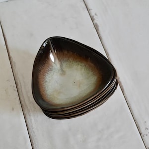 May include: A stack of five triangular ceramic bowls. The bowls have a gradient color scheme, transitioning from a dark brown rim to a light green center. The bowls are stacked on a white wooden surface.