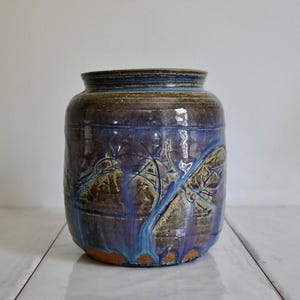 May include: A handcrafted ceramic vase with a wide, cylindrical shape. The vase features a textured surface with a dark blue and brown glaze, accented by lighter blue streaks and leaf-like patterns. The base has a reddish-brown hue.