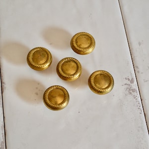 May include: Five circular brass knobs arranged in a cluster. Each knob features a smooth, slightly domed center surrounded by a raised, textured rope-like border. The metal has a muted, aged gold finish with subtle variations in tone and texture.