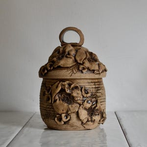 May include: A ceramic jar with a lid featuring a floral design. The jar is brown and has a textured surface. The lid has a ring handle.