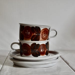 Peut inclure: Deux tasses en céramique blanche empilées sur deux soucoupes blanches assorties. Chaque tasse présente un motif répétitif de formes florales abstraites brun foncé et brun rougeâtre. Les tasses sont cylindriques avec une anse latérale.