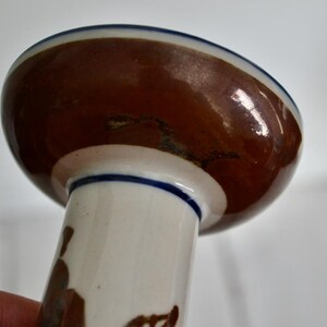 Puede incluir: Un candelero de cer&aacute;mica blanca con la parte superior esmaltada en marr&oacute;n y un borde azul. El candelero tiene una base ancha y un tallo alto y delgado.