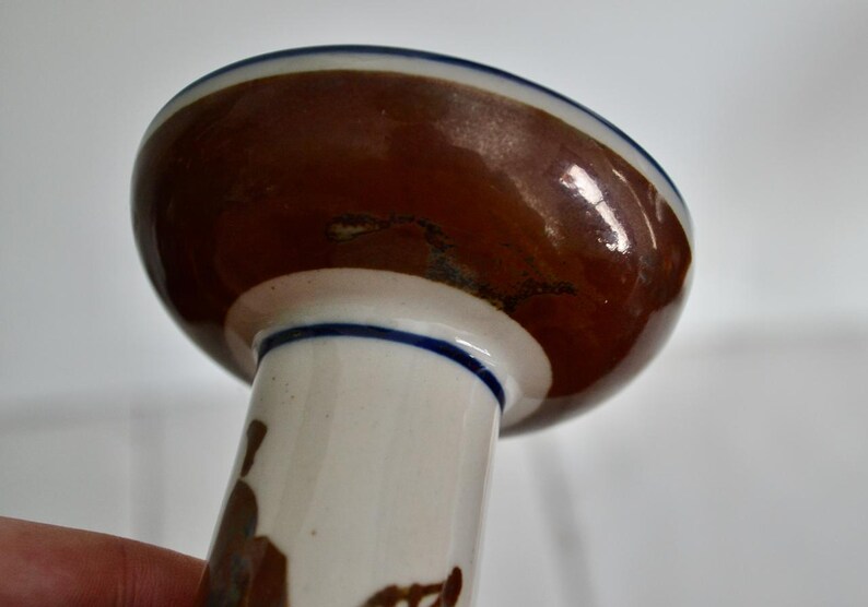 Puede incluir: Un candelero de cer&aacute;mica blanca con la parte superior esmaltada en marr&oacute;n y un borde azul. El candelero tiene una base ancha y un tallo alto y delgado.