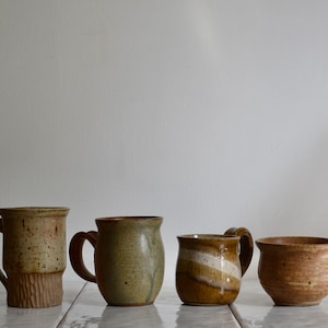 May include: Four ceramic mugs arranged in a row. The first is tall and speckled tan with a textured base. The second is a rounded olive green mug. The third is a brown mug with a white horizontal band. The fourth is a short, wide tan bowl-shaped mug.