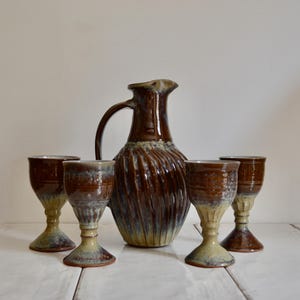 May include: A ceramic pitcher and four matching goblets featuring a glossy brown glaze that transitions into a muted green at the base. The pitcher has a ribbed, spiraling texture on its body, while the goblets have a smooth, stemmed design.