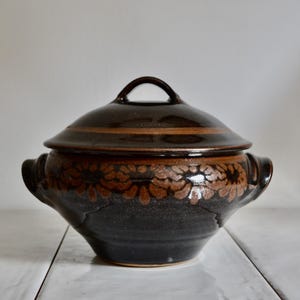 May include: A dark brown ceramic lidded casserole dish with two side handles. The body and lid feature a hand-painted band of light brown floral patterns. The glaze is glossy, and the vessel has a rounded shape with a tapered base and a knob handle.