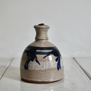 May include: A handmade ceramic bottle with a rounded base and a narrow neck. The bottle is beige with dark blue abstract brushstrokes. The bottle has a small, dark stopper.