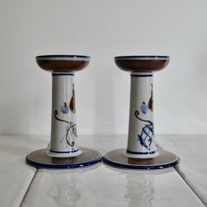 Puede incluir: Un par de candelabros de cer&aacute;mica blanca con dise&ntilde;os pintados en marr&oacute;n y azul. Los candelabros tienen una base ancha y una columna alta y delgada.