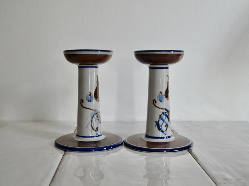 Puede incluir: Un par de candelabros de cer&aacute;mica blanca con dise&ntilde;os pintados en marr&oacute;n y azul. Los candelabros tienen una base ancha y una columna alta y delgada.