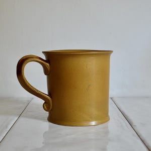 May include: Mustard-colored ceramic mug with a curved handle. The mug has a cylindrical shape with a slightly flared base and a smooth, matte finish. The handle has a decorative, curved design.