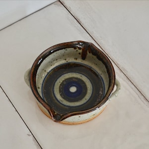 Peut inclure: Un petit plat rond en céramique avec un bord brun et un motif de cercles concentriques bleus et blancs. Le plat a deux petites poignées sur les côtés.