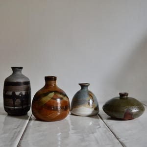 May include: Four handcrafted ceramic vases of varying shapes and sizes. The vases feature a range of colors, including brown, gray, blue, and green. The vases are displayed on a white, wooden surface.
