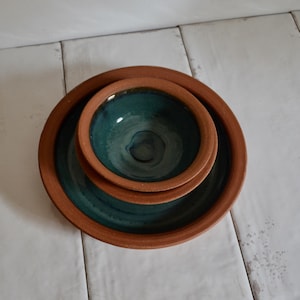 May include: A set of two nested ceramic dishes featuring a natural terracotta exterior and a deep teal glazed interior. The smaller bowl is centred within the larger plate, both displaying a smooth, rounded rim and a contrasting colour palette.