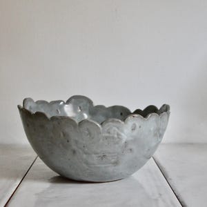 May include: A grey ceramic bowl with a scalloped edge. The bowl has a textured surface and is placed on a white wooden surface. The bowl is empty and looks handmade.