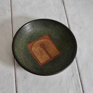 May include: A handmade ceramic bowl with a dark green interior and a black rim. The bowl features a square, tan-coloured design in the centre. The bowl is round and sits on a white surface.