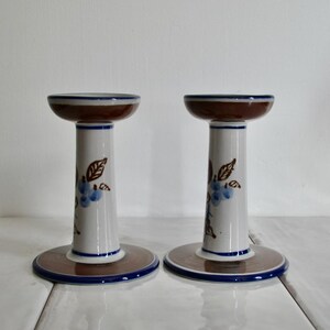 Puede incluir: Un par de candelabros de cer&aacute;mica blanca con dise&ntilde;os florales marrones y azules. Los candelabros tienen una base ancha y una parte superior estrecha.