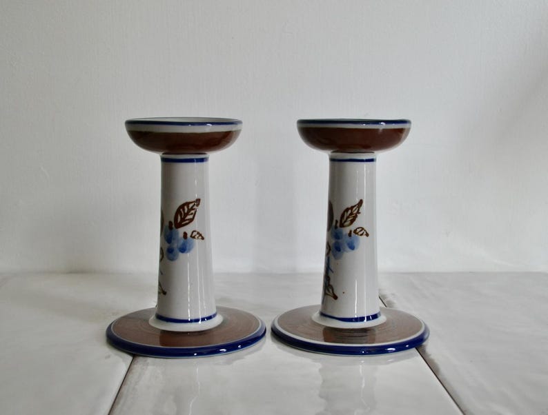 Puede incluir: Un par de candelabros de cer&aacute;mica blanca con dise&ntilde;os florales marrones y azules. Los candelabros tienen una base ancha y una parte superior estrecha.