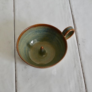 May include: A handmade ceramic apple baker with a handle. The bowl is glazed in shades of green and blue, with a brown rim and a central spike for holding an apple. The baker is designed for baking apples and other fruits.