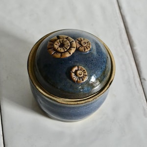 May include: A handmade ceramic jar with a lid. The jar is a deep blue with a speckled glaze, and the lid features three raised, flower-like designs in a contrasting tan colour. The jar has a round shape and a matching rim.